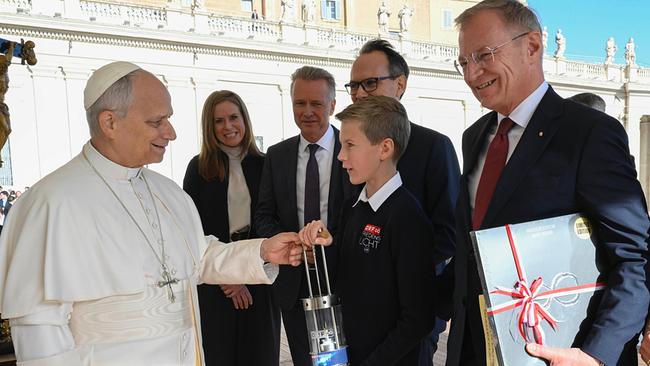 Papst Leo XIV. nimmt am Petersplatz das „ORF-Friedenslicht aus Bethlehem“ von Friedenslichtkind Florian Mitter entgegen, während mehrere Begleitpersonen danebenstehen und ein Mann ein Geschenkpaket hält.
