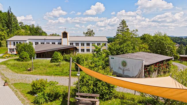 Das Haus der Stille: eine grüne Anlage mit mehreren Gebäuden, darunter ein größeres Haus mit Kreuz auf dem Dach, umgeben von Bäumen, Wiesen und einem Sonnensegel im Vordergrund.