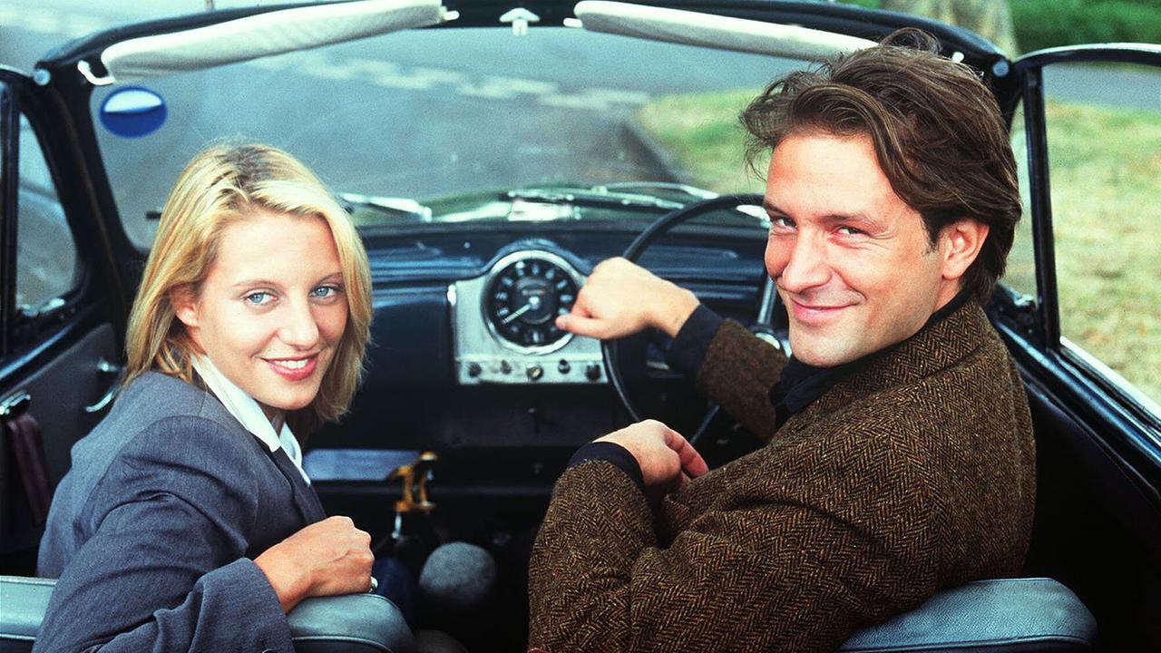 Eine Frau und ein Mann sitzen in einem kleinen Sportwagen und blicken durch das geöffnete Verdeck nach hinten und lächeln freundlich