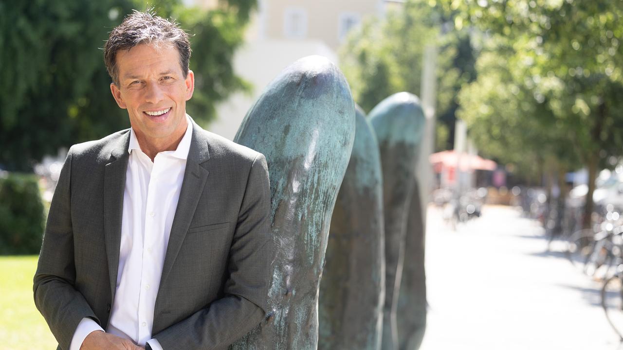 © ORF/Landesstudio Salzburg/wildbild Moderator Martin Ferdiny im grauen Anzug und weißen Hemd steht im Freien neben einer abstrakten Skulptur an einem sonnigen Tag.