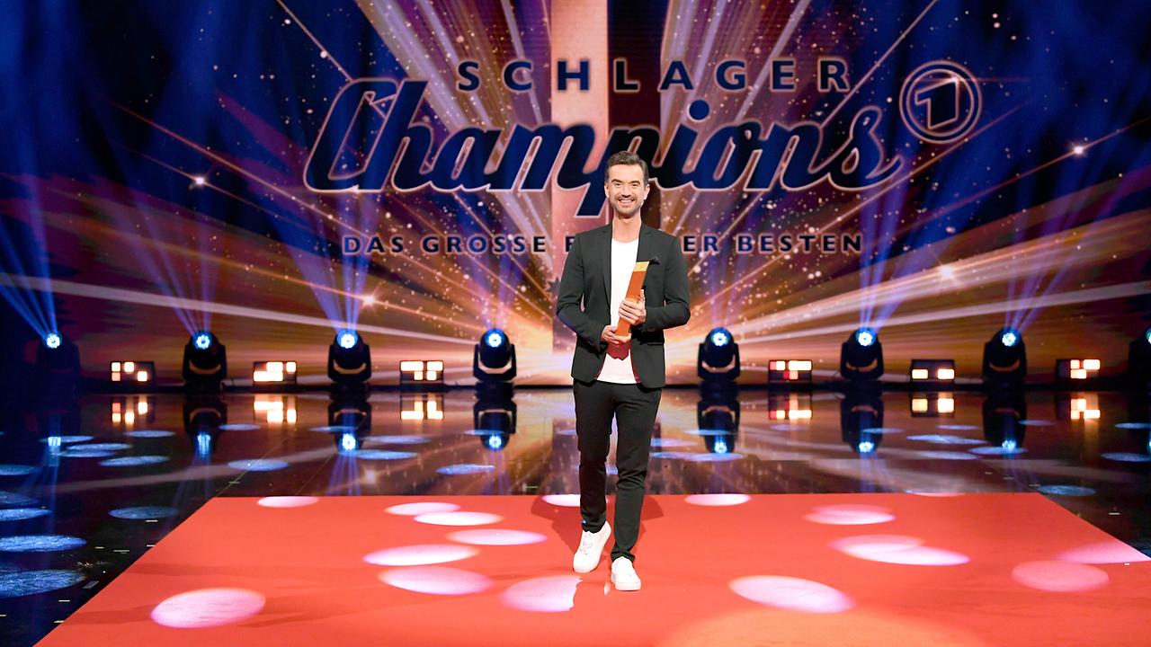 Florian Silbereisen steht auf einem rot beleuchteten Show‑Boden und hält eine Auszeichnung auf der  bunt ausgeleuchtete Bühnenbild zu „Schlagerchampions – Das große Fest der Besten“.