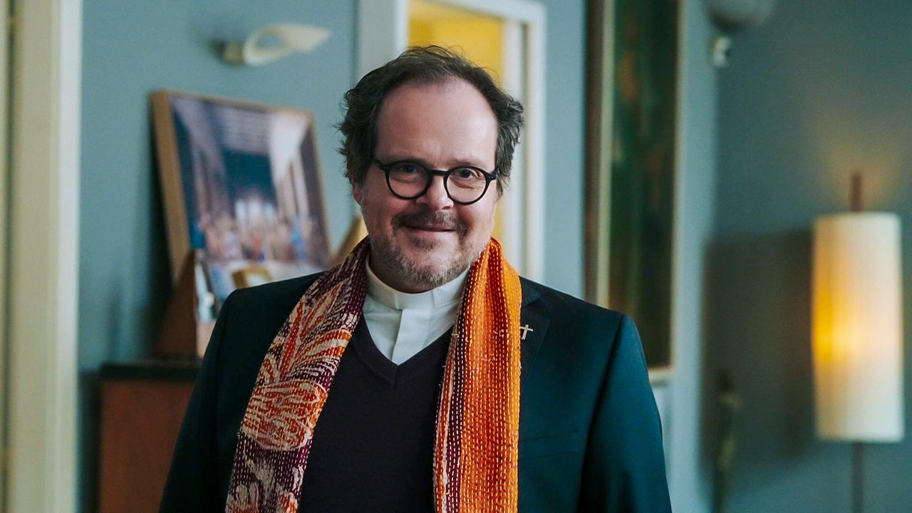 © ORF/BR/Cult Film Edgar in dunkler Kleidung mit farbigem Schal und Kreuz-Anstecker steht in einem Wohnraum.