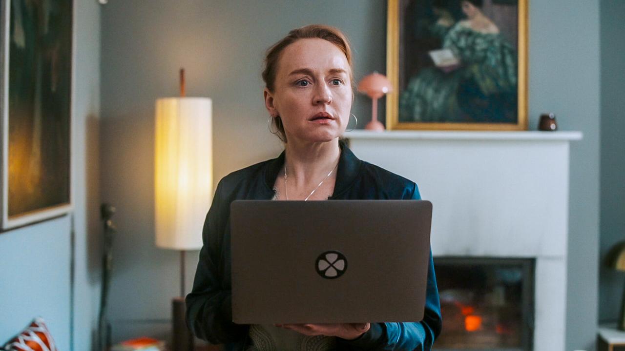 © ORF/BR/Cult Film Balbina steht mit einem Laptop vor einem Kamin im Wohnzimmer und blickt nach vorne.