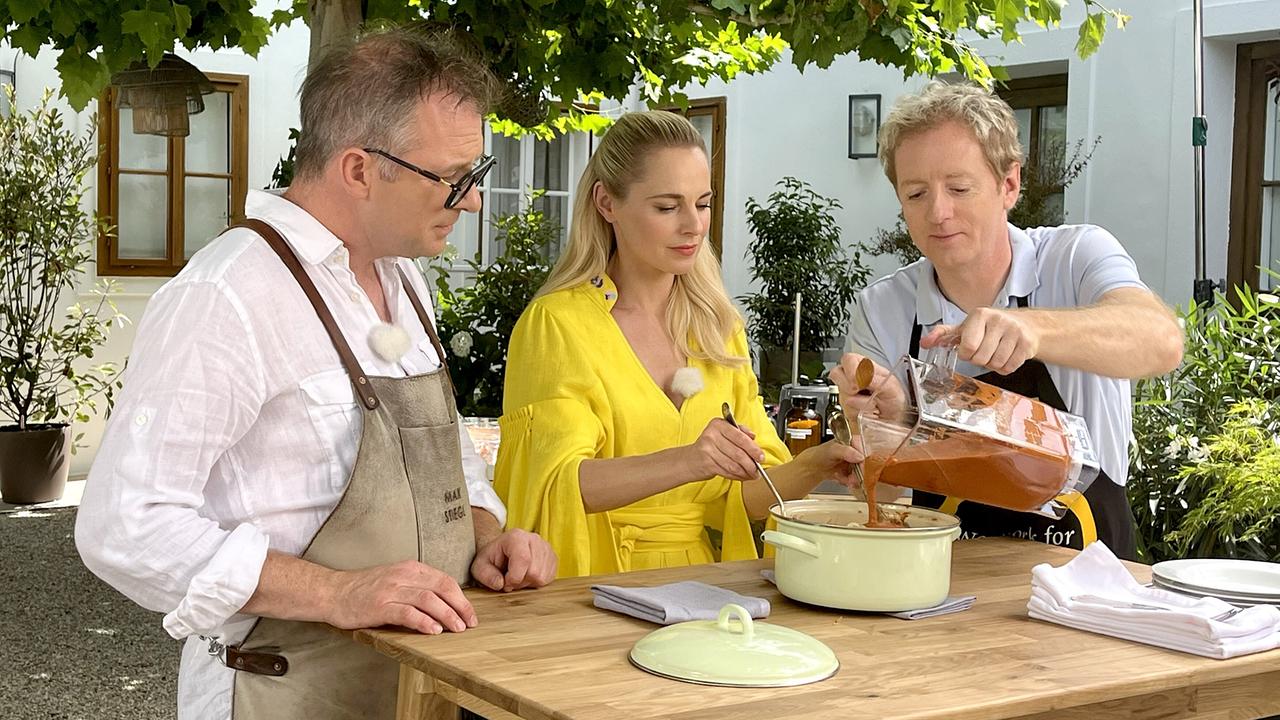 Max Stiegl, Silvia Schneider und Fritz Treiber an einem Holztisch im Freien, Fritz gießt Pesto über die Pasta