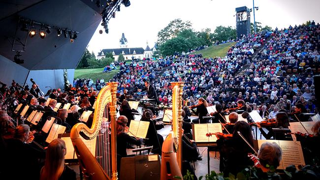 © ORF/Roman Zach-Kiesling Ein Orchester spielt ein Konzert auf einer Freiluftbühne im Wolkenturm Grafenegg vor einem voll besetzten Amphitheater in parkähnlicher Umgebung.