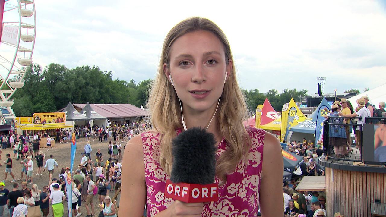 © ORF Moderatorin Isabella Köck steht mit ORF-Mikro auf einem Festivalgelände vor Publikum, Riesenrad und Bühne beim Woodstock.