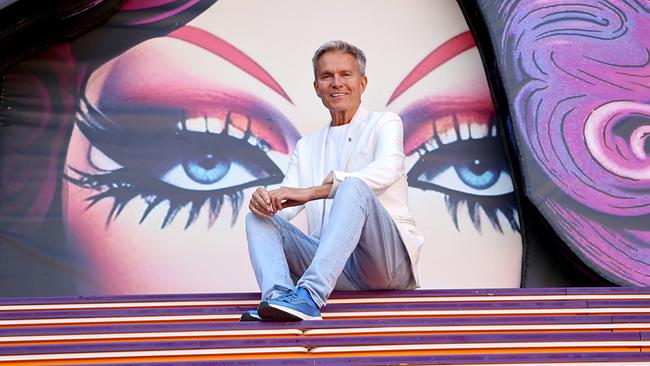 © ORF/Landesstudio Burgenland/Markus Fuchs Moderator Alfons Haider sitzt lächelnd auf bunten Stufen vor einer riesigen Darstellung geschminkter Augen auf der Bühne von „Ein Käfig voller Narren“ auf der Seebühne Mörbisch.