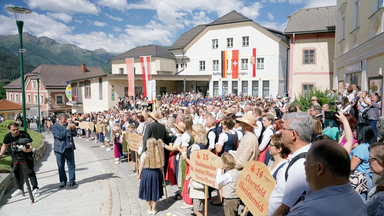 Zahlreiche Trachtenchöre und Besucherinnen und Besucher versammeln sich bei strahlendem Wetter auf einem Dorfplatz in den Bergen beim Chorfestival "Feuer & Stimme" in St. Michael.