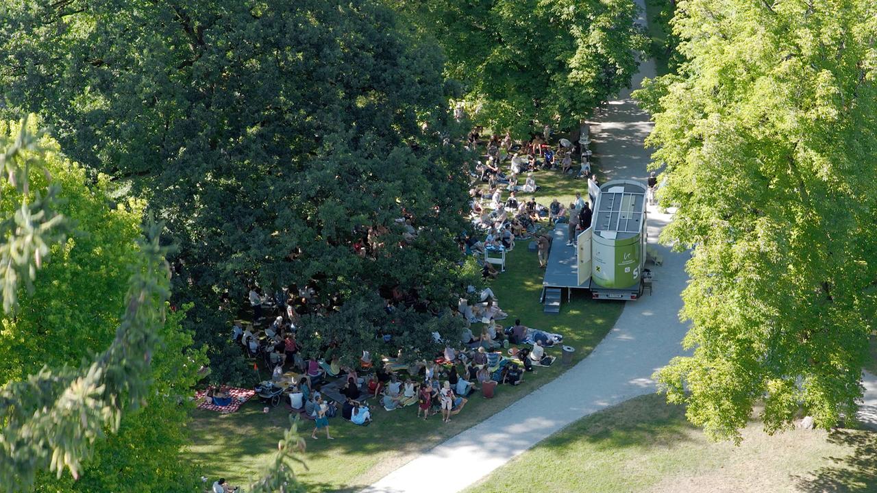 Menschen sitzen auf Decken und Campingstühlen im Schatten eines großen Baums und verfolgen eine Vorstellung auf einer mobilen Bühne beim Straßentheater Hellbrunn.