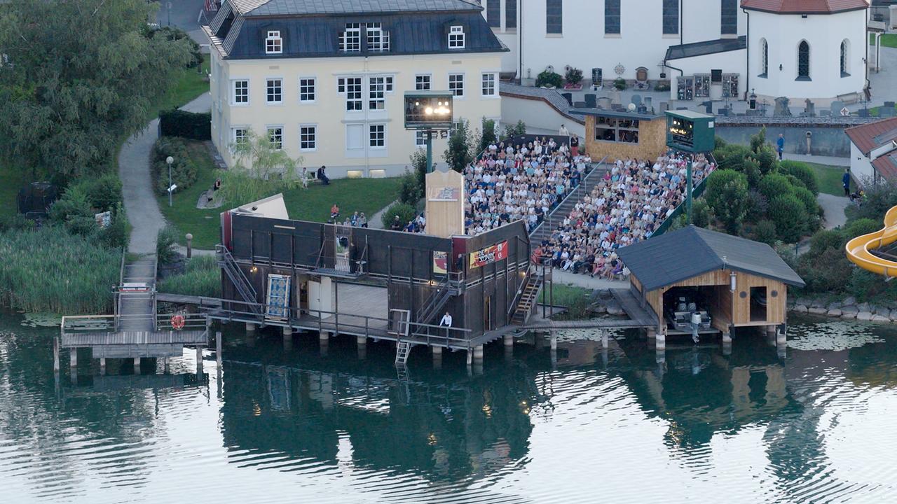 Eine Luftaufnahme zeigt die Seebühne Seeham neben Kirche und Wohnhaus sowie ein voll besetztes Freilufttheater.