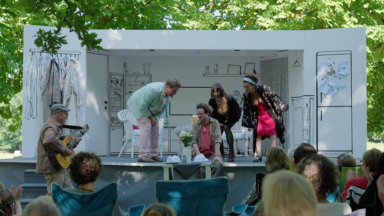 Eine kleine Freilufttheaterbühne im Park beim Straßentheater Hellbrunn zeigt eine Wohnzimmerkulisse, vor der mehrere Schauspielerinnen und Schauspieler auftreten.