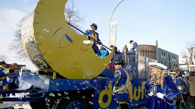 Ein Faschingswagen mit einer großen gelben Mondfigur in Bewegung, umgeben von kostümierten Teilnehmern in Blau-Gelb, während Seifenblasen durch die Luft fliegen.