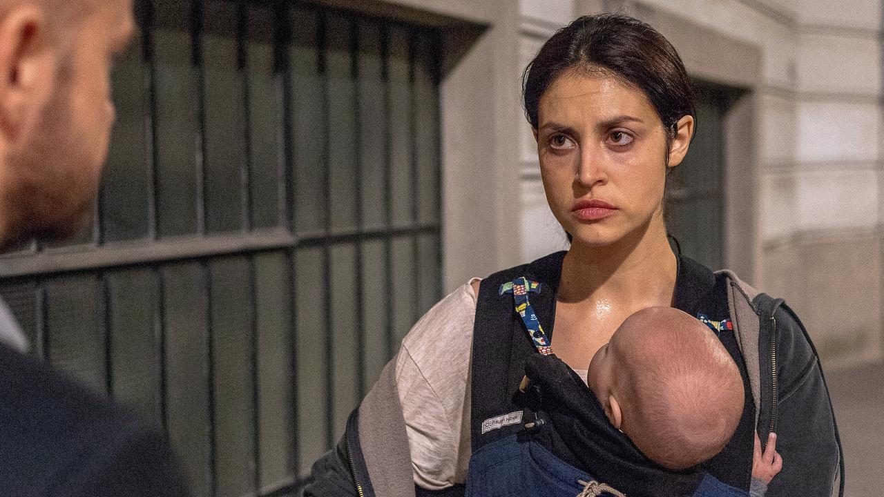 Leila Nymann steht im Freien und trägt ein Baby in einer Trage vor der Brust, während sie mit jemandem spricht.