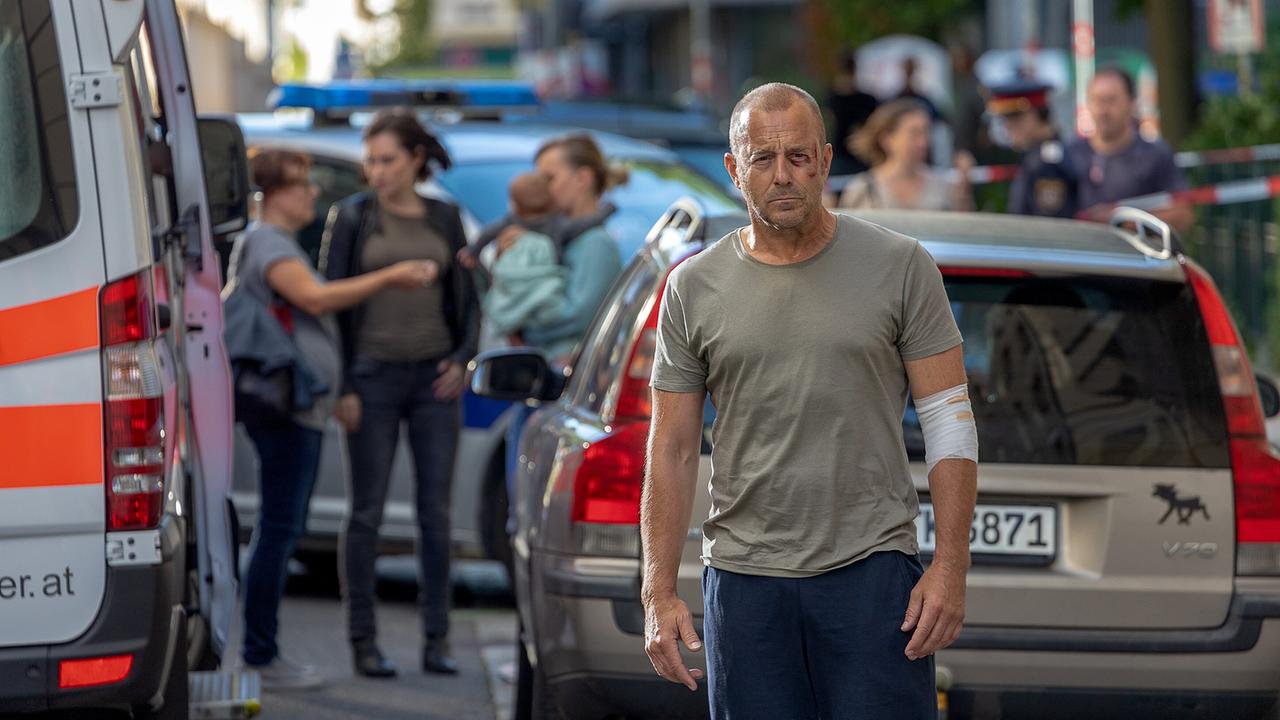 Richard Brock mit bandagiertem Arm steht auf einer Straße, im Hintergrund sind ein Rettungsfahrzeug, ein Polizeiauto und mehrere Menschen zu sehen.