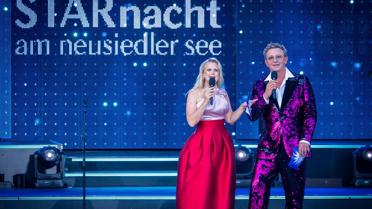 Auf einer beleuchteten Open-Air-Bühne moderieren Barbara Schöneberger und Hans Sigl  vor dem Schriftzug „STARnacht am Neusiedler See“.