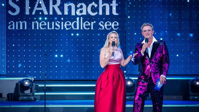 Auf einer beleuchteten Open-Air-Bühne moderieren Barbara Schöneberger und Hans Sigl  vor dem Schriftzug „STARnacht am Neusiedler See“.