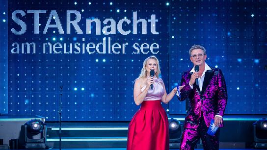 Auf einer beleuchteten Open-Air-Bühne moderieren Barbara Schöneberger und Hans Sigl  vor dem Schriftzug „STARnacht am Neusiedler See“.