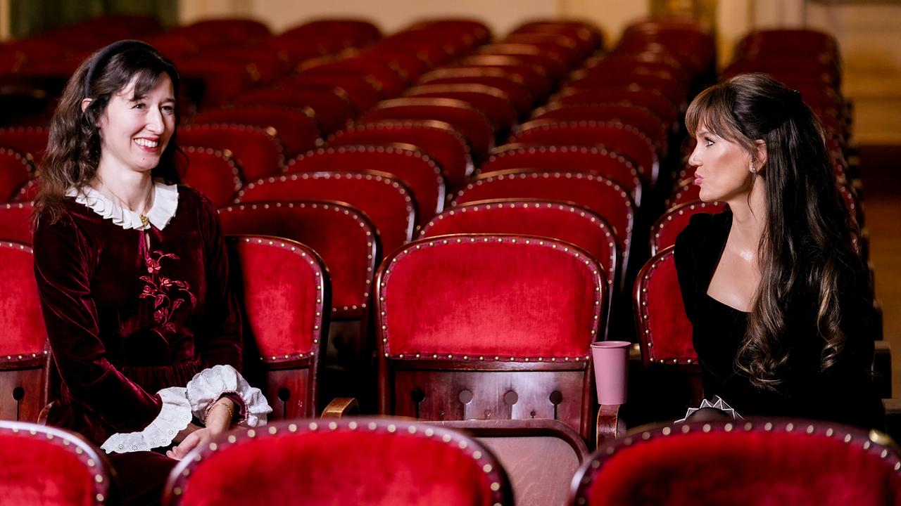 Maddalena del Gobbo und Leona König  sitzen mit Abstand in einem leeren Theatersaal auf roten Polstersitzen und unterhalten sich.