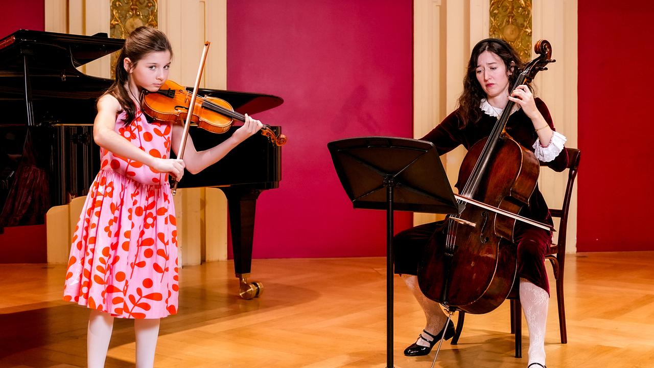Eloisa Aubert an der Violine und Maddalena del Gobbo am  Cello musizieren gemeinsam in einem prunkvollen Raum mit einem Flügel im Hintergrund.
