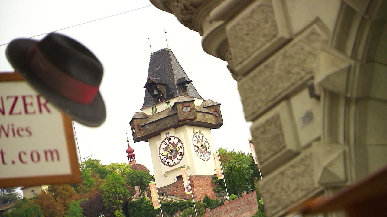 © ORF/Landesstudio Steiermark Der Grazer Uhrturm ist aus der Perspektive zwischen einem Hut und einem steinernen Gebäudeelement zu sehen.