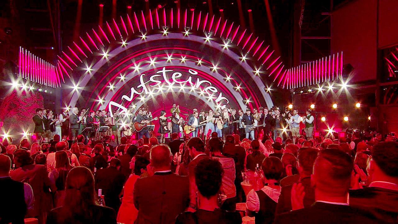 © ORF/Landesstudio Steiermark Ein großes Ensemble feiert unter rotem Licht auf der "Aufsteirern"-Bühne vor einem dicht gefüllten Saal.