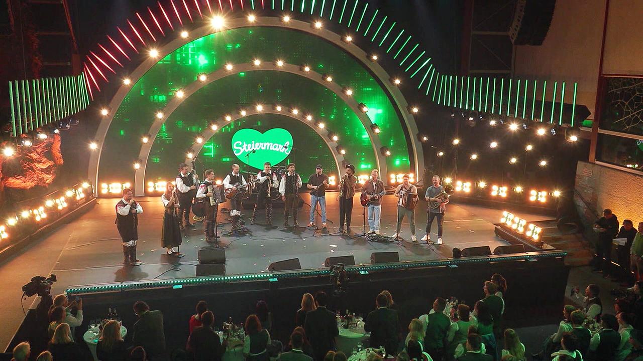 © ORF/Landesstudio Steiermark Zwölf Musikerinnen und Musiker stehen vor einem Publikum auf einer groß ausgeleuchteten Bühne mit grünem Herz mit einem „Steiermark“-Schriftzug in der Mitte.