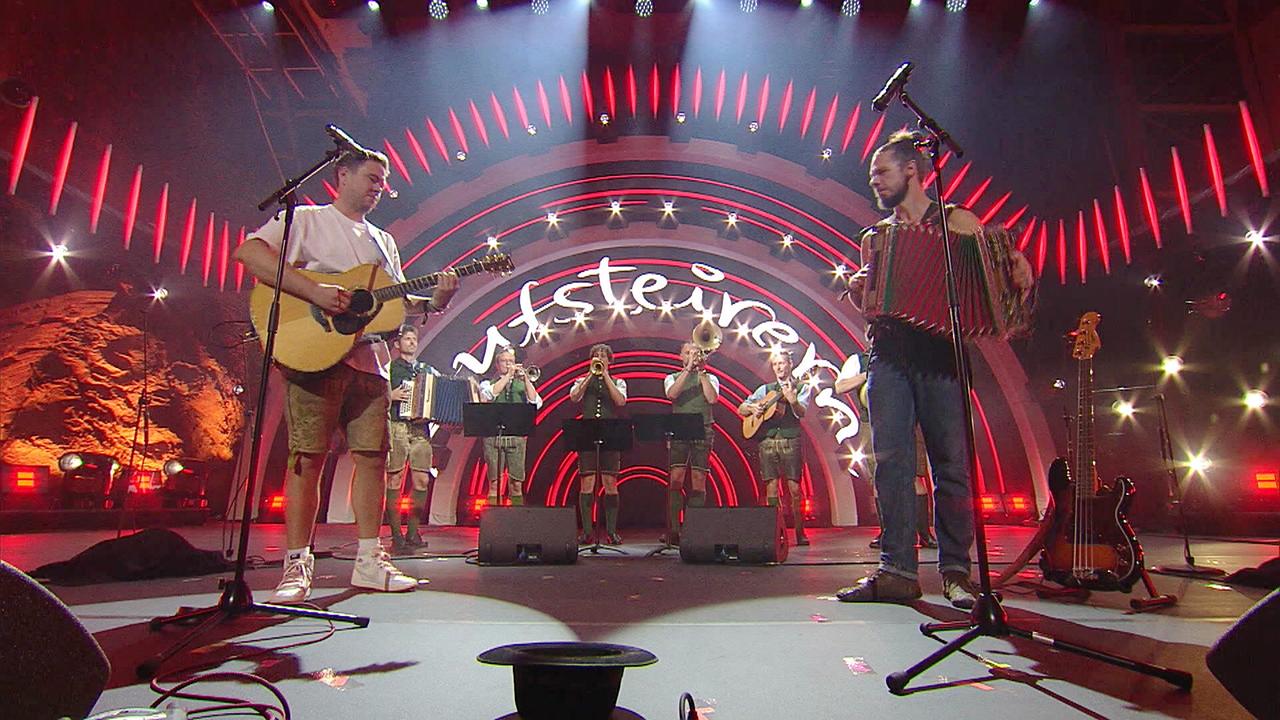 © ORF/Landesstudio Steiermark Eine Musikgruppe spielt mit Gitarren, Akkordeon und Blasinstrumenten auf der Bühne der „Aufsteirern“-Veranstaltung.