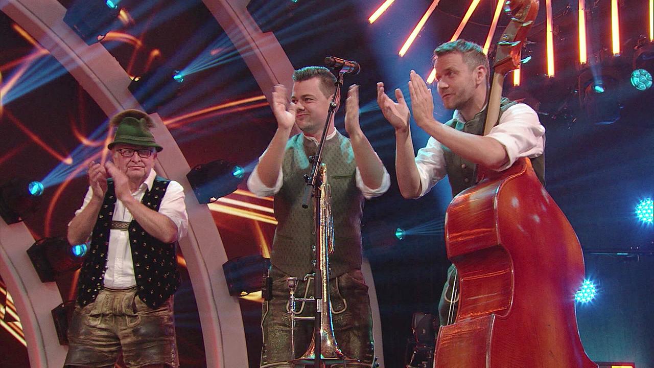 © ORF/Landesstudio Steiermark Drei Musiker in Tracht applaudieren auf einer bunt beleuchteten Bühne, vor ihnen stehen Mikrofon, Posaune und Kontrabass.