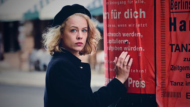 Stella steht vor einem roten Plakat an einer Litfaßsäule auf einer Straße.