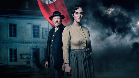 Josef Hader und Verena Altenberger vor einem mit Wolken verhangenem Haus und einer Fahne