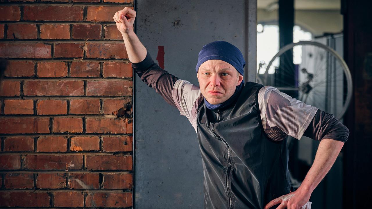 Eddi, der studiert und zu den Velopunks gehört, lehnt mit erhobenem Arm an einer Backsteinwand in einem industriellen Innenraum.