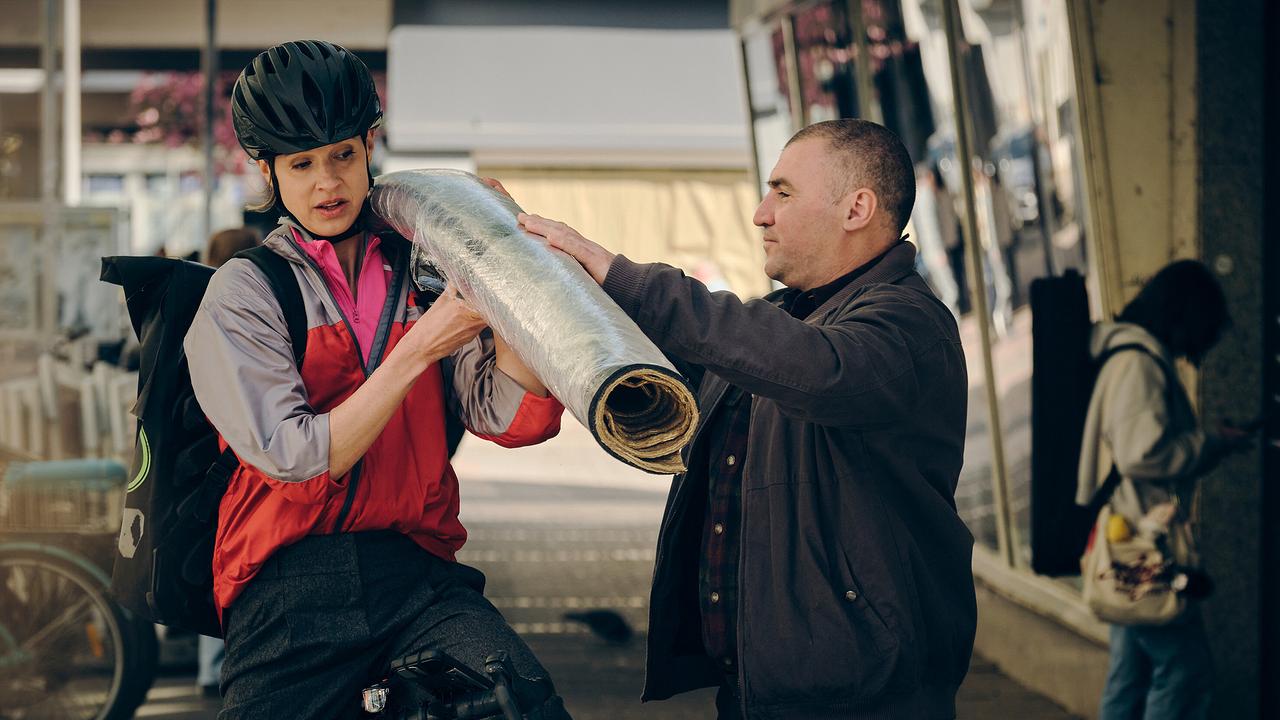 In einer überdachten Halle übergibt ein Mann der Radfahrerin Johanna Stern eine große, zusammengerollte Matte.