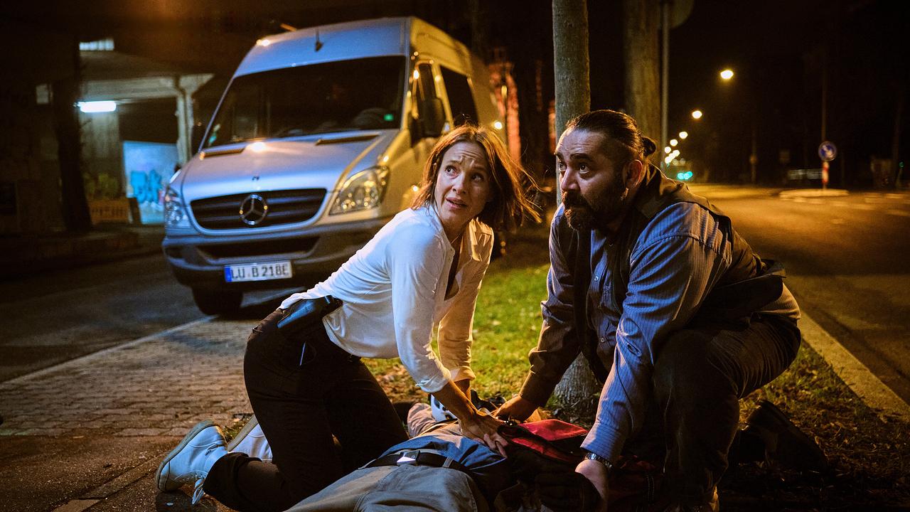 Johanna und Dr. Özcan knien nachts am Straßenrand und leisten Erste Hilfe bei einem am Boden liegenden Fahrradkurier, nachdem er überfahren wurde.