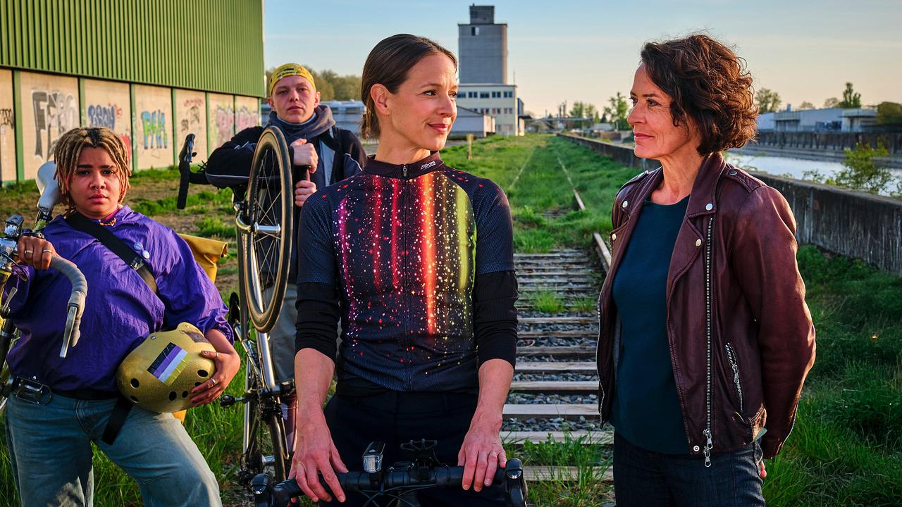 Zwei Fahrradfahrer von Velopunks, Johanna Stern, die undercover ermittelt, und Lena Odenthal stehen neisammen auf stillgelegten Gleisen in einem industriellen Umfeld neben dem Rheinufer.