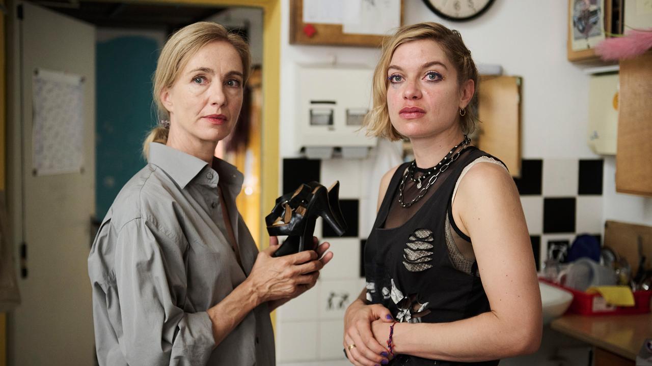 In einer Werkstatt hält Gina Rohland ein Paar schwarze Schuhe, während Garderobiere Ria Jäger danebensteht. Beide schauen in eine Richtung.