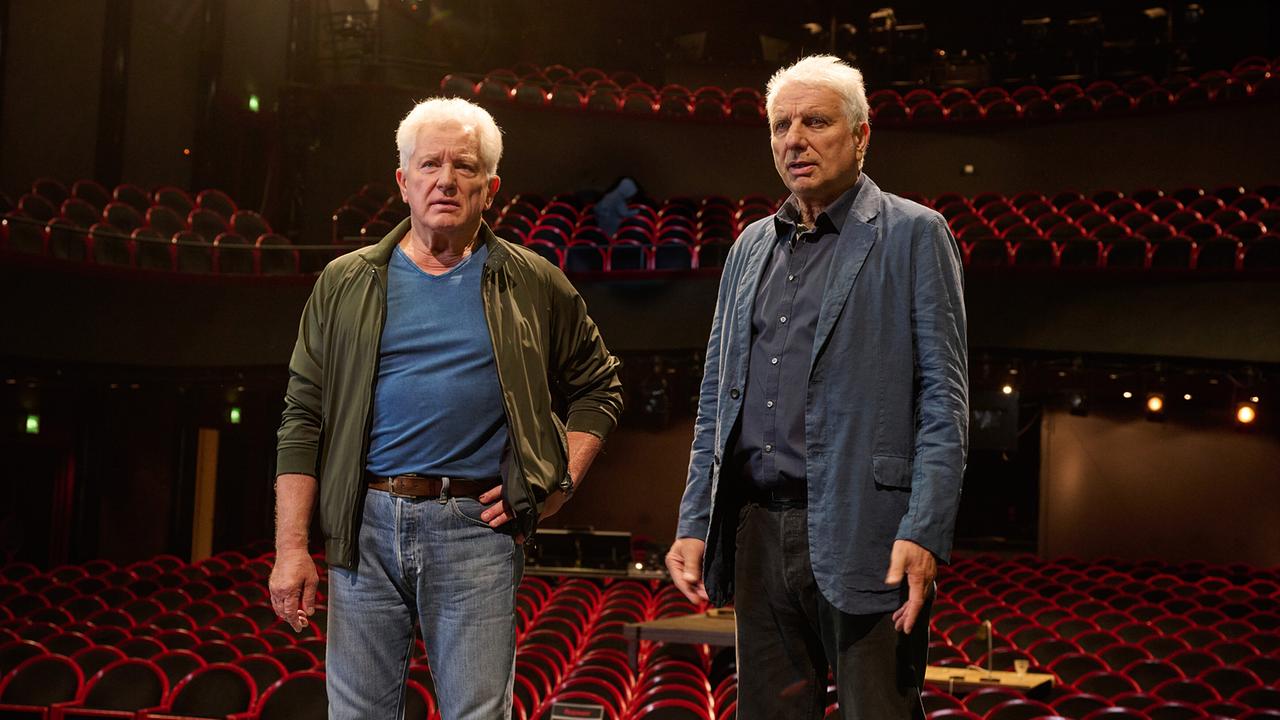 Kriminalhauptkommissar Ivo Batic und Kriminalhauptkommissar Leitmayr stehen auf einer Bühne vor einem Zuschauerraum mit roten Sitzen.