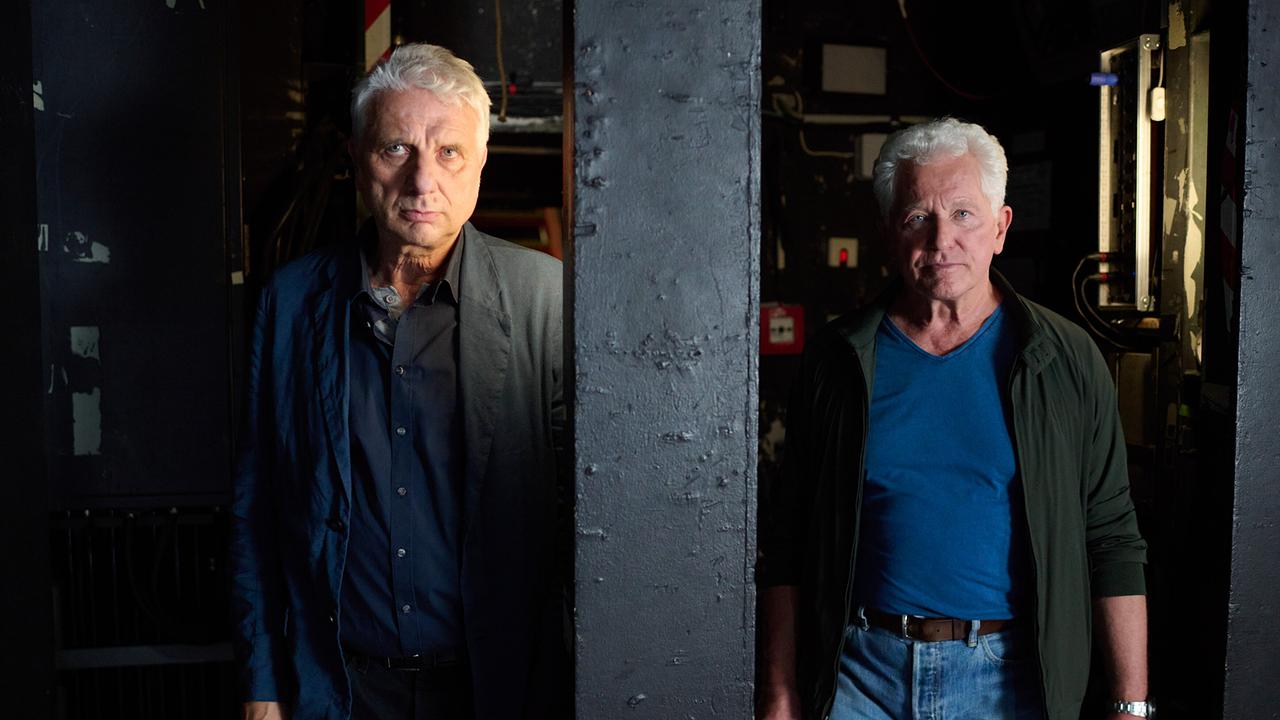 Kriminalhauptkommissar Leitmayr und Kriminalhauptkommissar Ivo Batic  getrennt durch einen Pfeiler beobachten in einem dunklen Backstage-Bereich das Geschehen auf der Bühne.