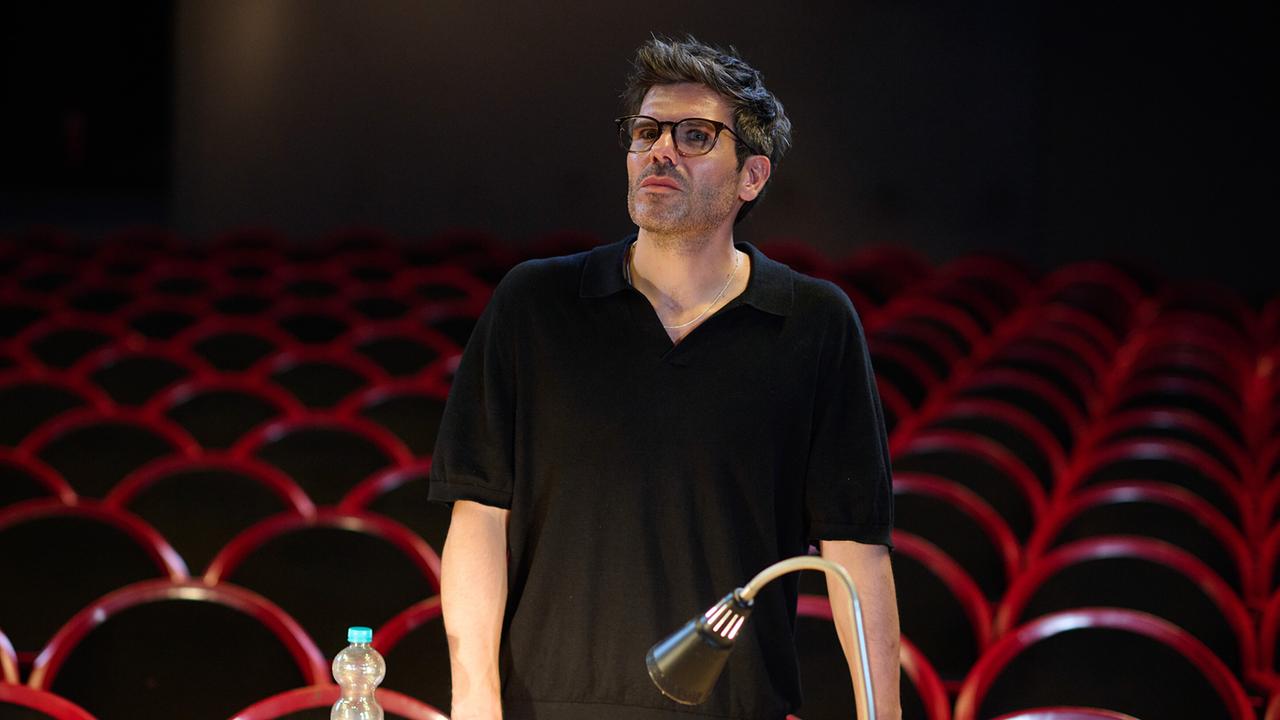 Regisseur Akim Birol steht im Theater vor leeren Reihen roter Sitzplätze an einem Pult mit Lampe und Wasserflasche und ist mit Carls Spiel nicht zufrieden.