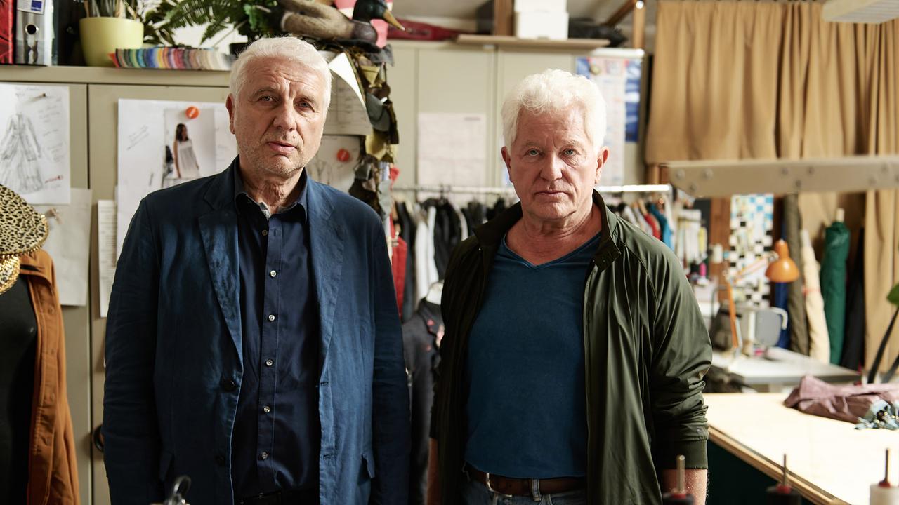 Kriminalhauptkommissar Leitmayr und sein Kollege Kriminalhauptkommissar Ivo Batic stehen in einer Werkstatt zwischen Stoffen, Schnitten und Nähutensilien, um Gina und Ria zu befragen.