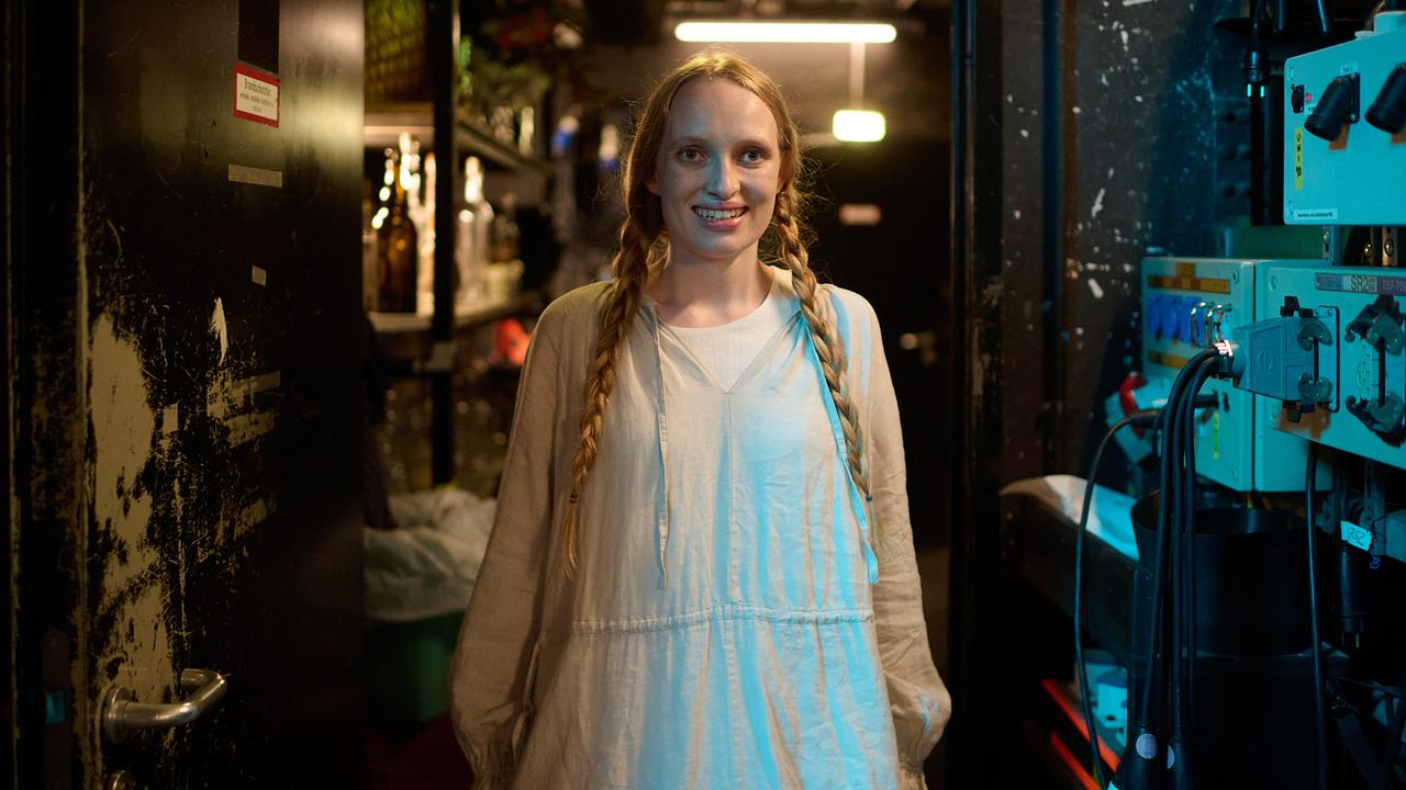 Stella mit zwei Zöpfen steht lächelnd in einem engen Technik-/Backstage-Gang zwischen Geräten und Regalen.