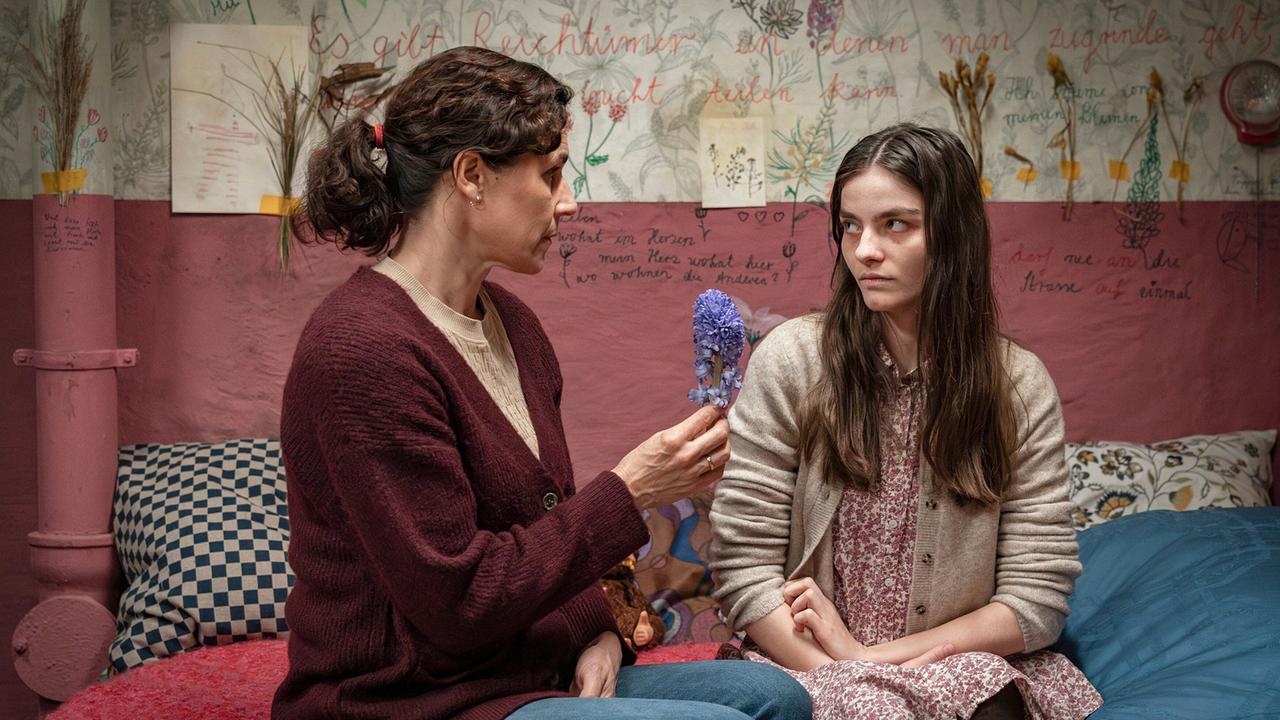 In einem bunt gestalteten Zimmer vor einer angekritzelten Wand sitzt Amanda mit ihrer Mutter, die eine Blume in der Hand hält, am Bett.