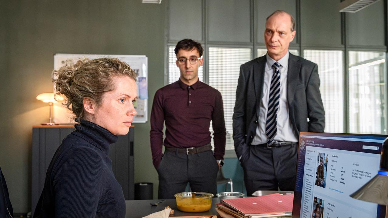 Leonie Winkler, Philipp Laupheimer und Peter Schnabel sitzen und stehen  um einen Schreibtisch im Kommissariat und arbeiten am aktuellen Fall.