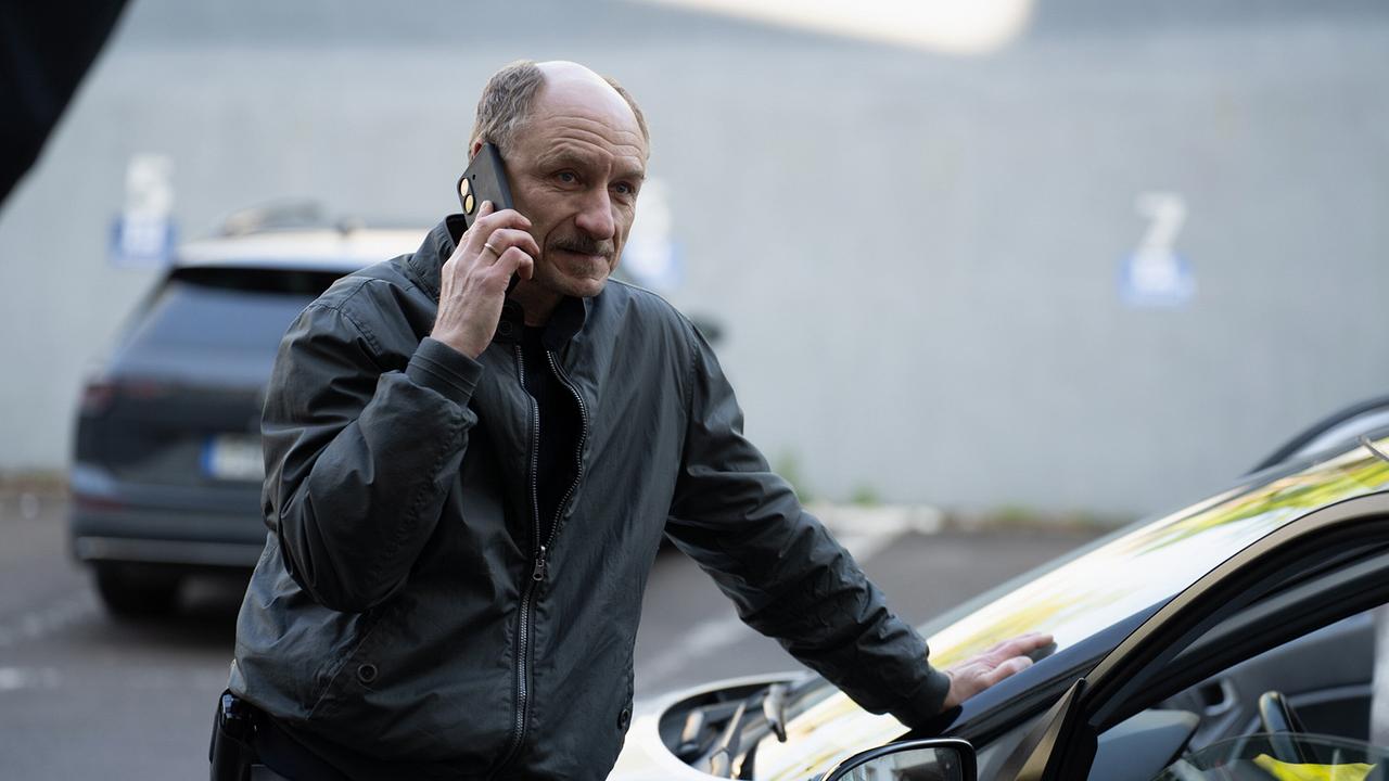 Christian Möller telefoniert neben einem geparkten Auto auf einem Parkplatz.