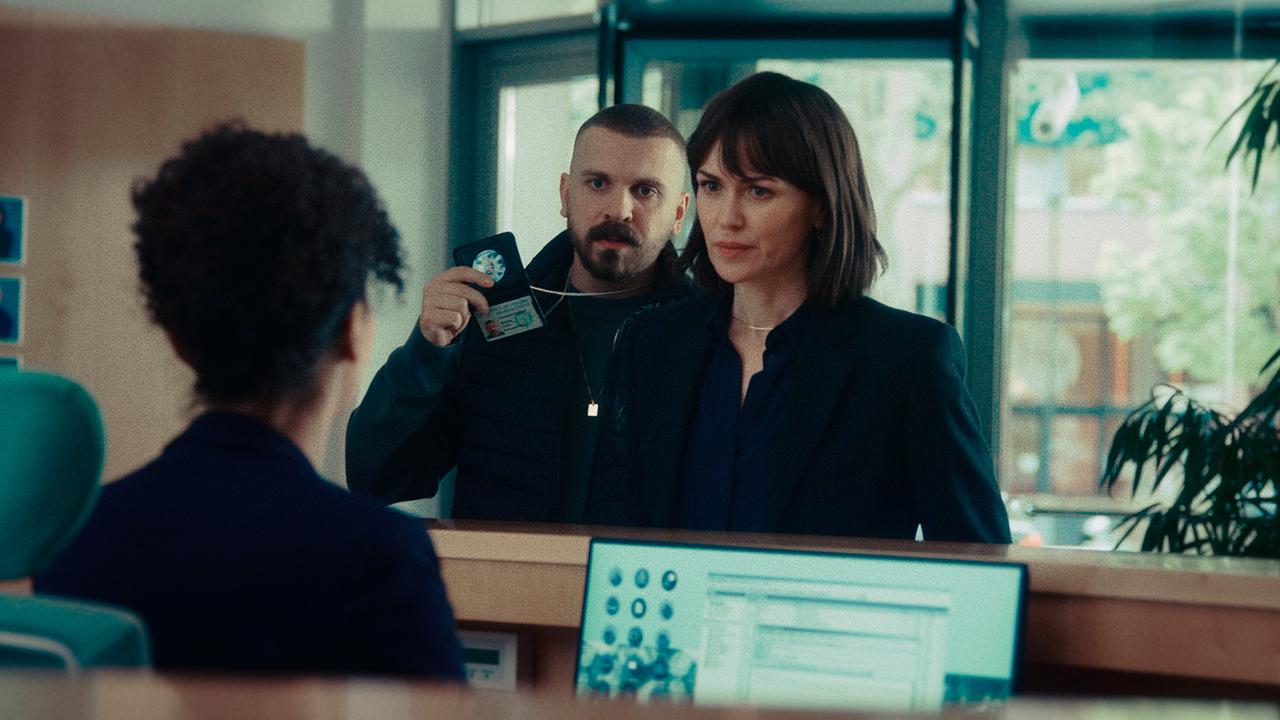 An einem Schalter stehen Maryam Azadi und Hamza Kulina, der seinen Dienstausweis zeigt, während eine Mitarbeiterin dahinter sitzt.