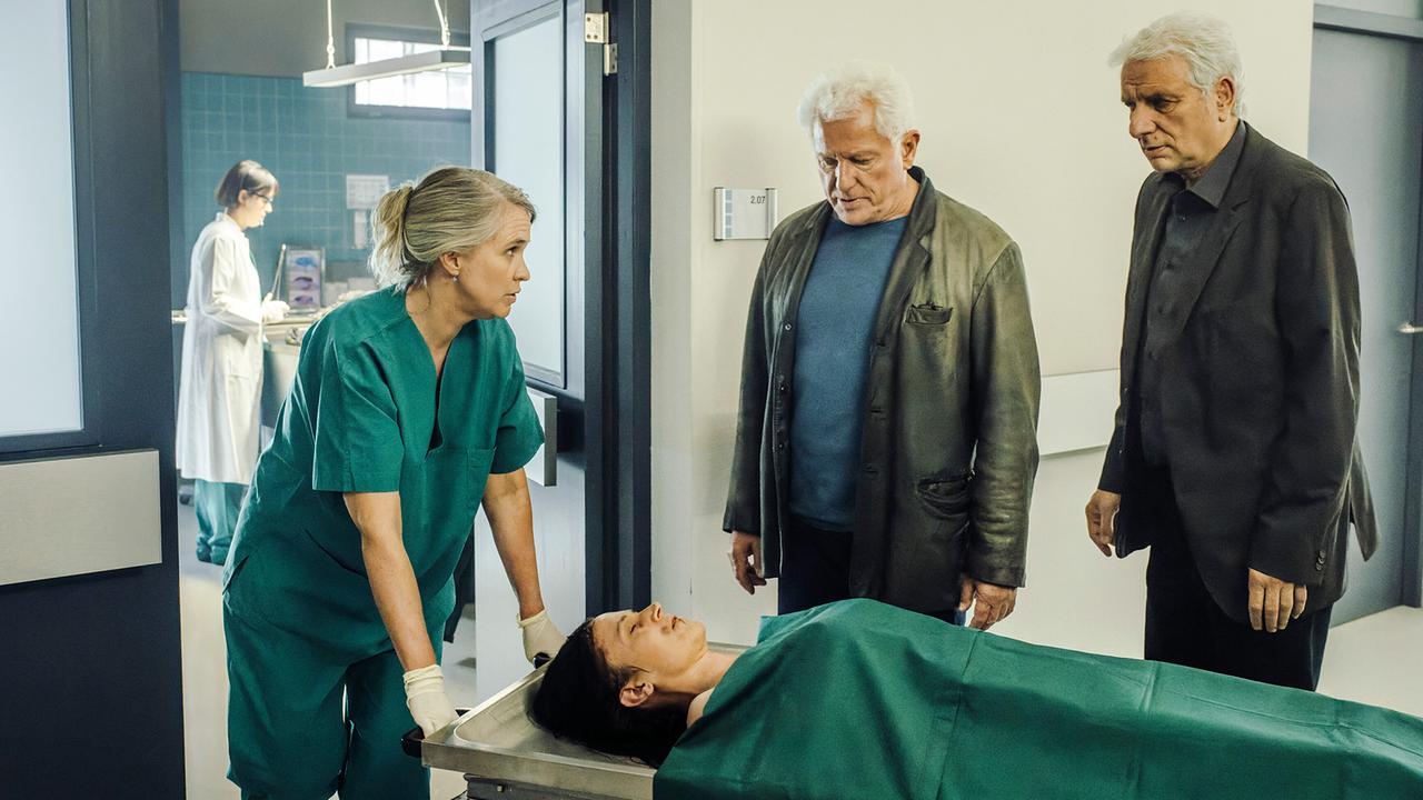 Eine Assistentin der Rechtsmedizin zeigt Ivo Batic und Franz Leitmayr  auf einer Trage eine weibliche Captagon-Leiche in einem Flur.