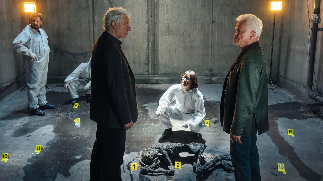 Kriminalhauptkommissar Franz Leitmayr, Rechtsmedizinerin Dr. Roswitha Schwarz-Fischer, Kriminalhauptkommissar Ivo Batic und weitere Kollegen stehen am Tatort mit der verkohlten Leiche im Lüftungsschacht unter dem St. Quirin Platz.