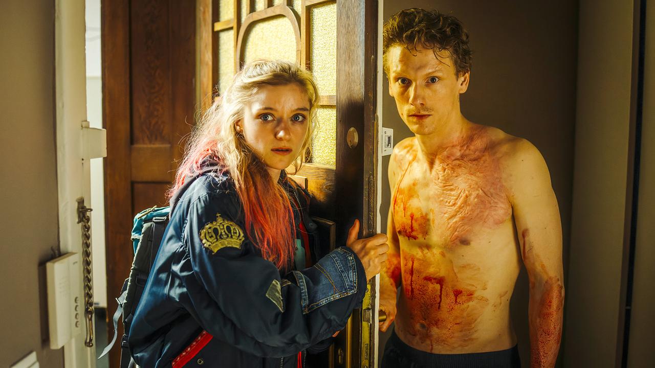 Kara Roth öffnet eine Wohnungstür, hinter der der verletzte, blutverschmierte Mike Werner mit nacktem Oberkörper steht.