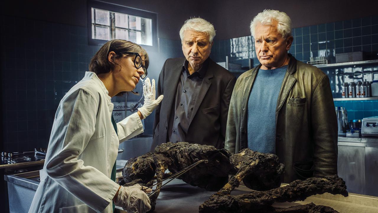 Die Rechtsmedizinerin Dr. Roswitha Schwarz-Fischer steht vor der verkohlten Leiche auf einem Edelstahltisch im forensischen Labor und spricht mit  Franz Leitmayr und Ivo Batic.