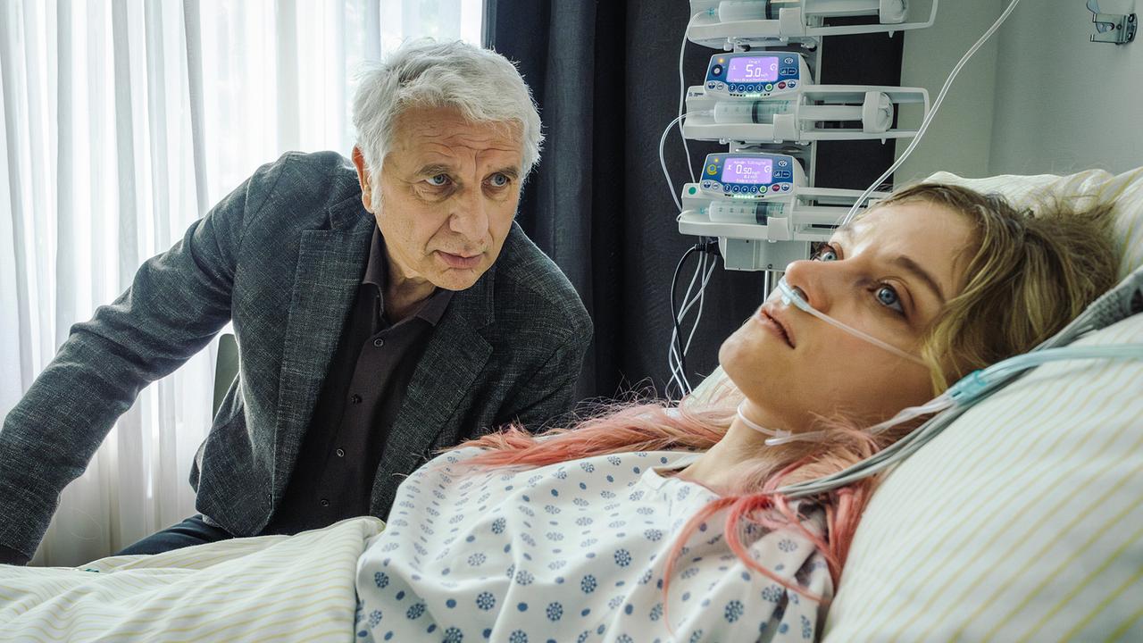 Franz Leitmayr  sitzt am Bett von Kara Roth in einem Krankenhauszimmer mit medizinischen Geräten im Hintergrund und versucht, an Informationen zu kommen.