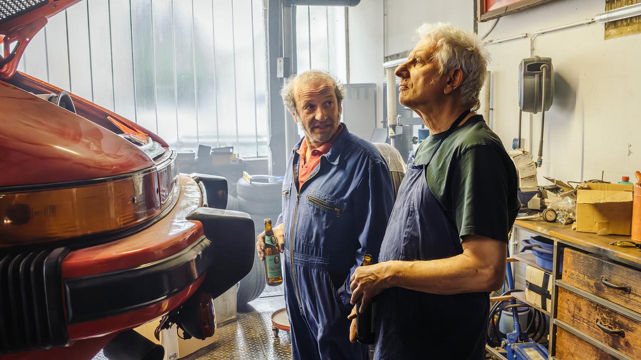 Mechaniker Detlef und Franz Leitmayr stehen in einer Werkstatt neben einem angehobenen Auto und halten eine Flasche Bier in der Hand.